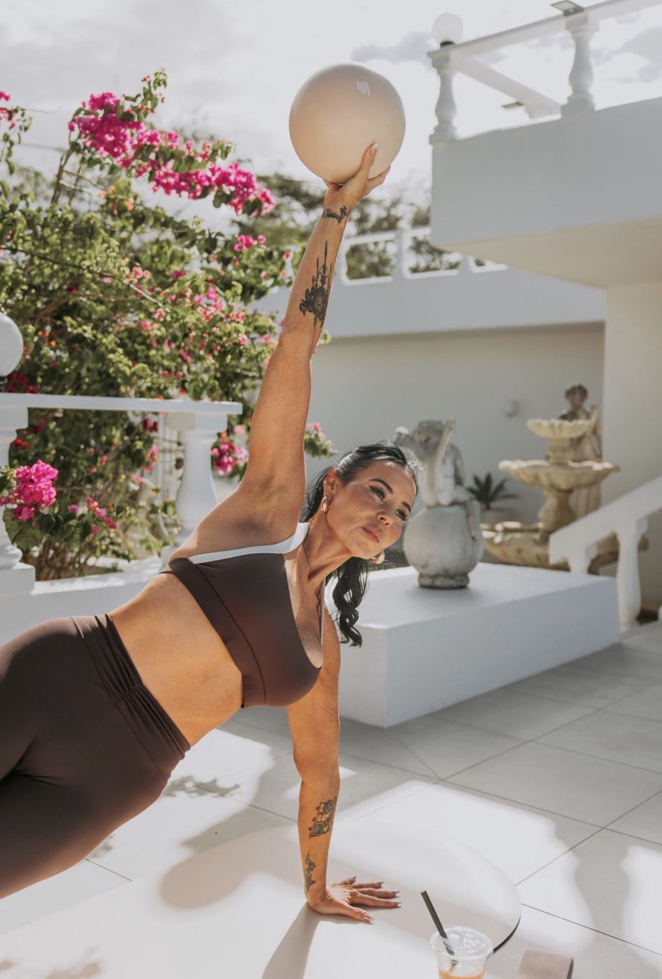 A UNLMTD client in a side plank holding a ball overhead beside pink bougainvillea